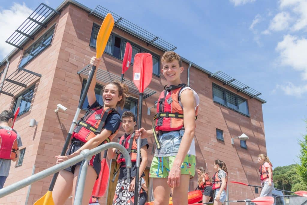 three students holding oars are happily standing wearing lifejackets outside the specialist watersports training centre in the background to do some canoeing