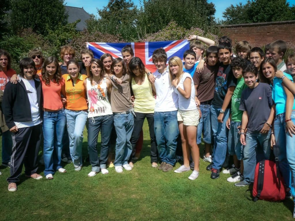 Spanish school group hugging each other and holding a UK flag up behind them as they smile at the camera in the garden of the Isca School of English