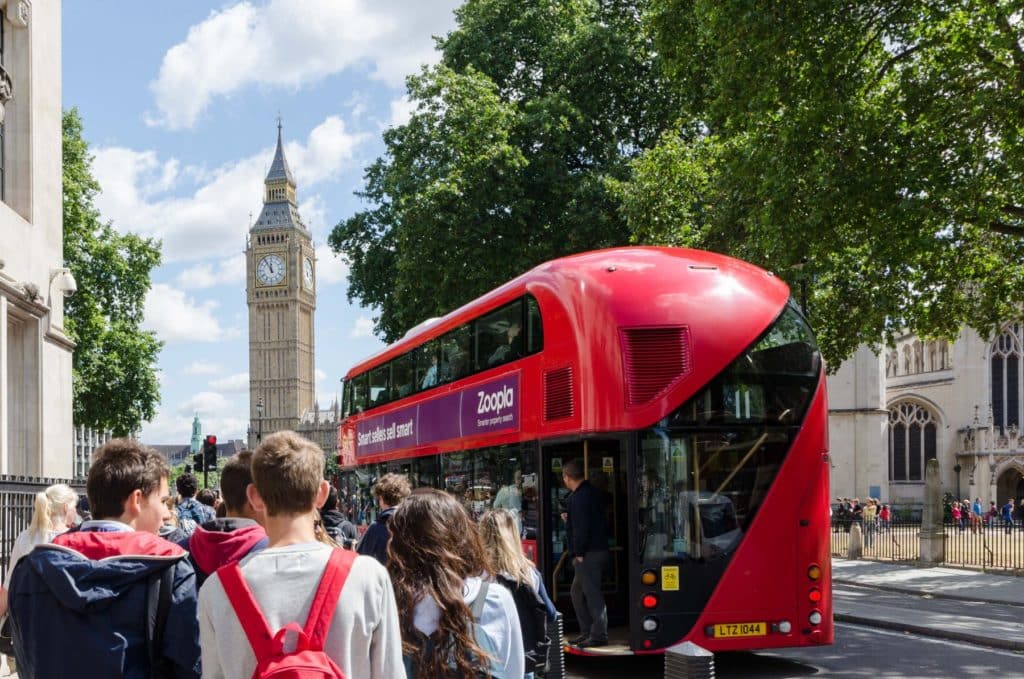 International students from behind walking into Trafalgar Square with a new red double decker bus on the road and Big Ben in the back ground. The side entrance to Westminister Abbey and tourists entering can also be seen
