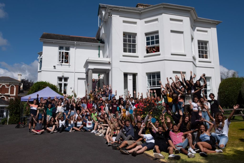 large group of waving students sitting and standing with their teachers in the front garden of the Isca School of English in Exeter. The school is a white georgian building and is behind the students