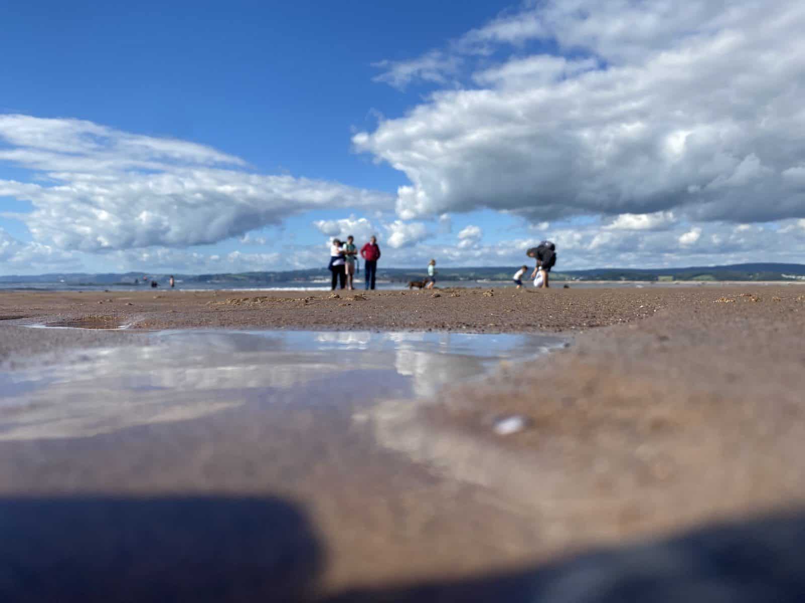 close up of water in the sand at Exmouth beach with a family playing in the background on a sunny day