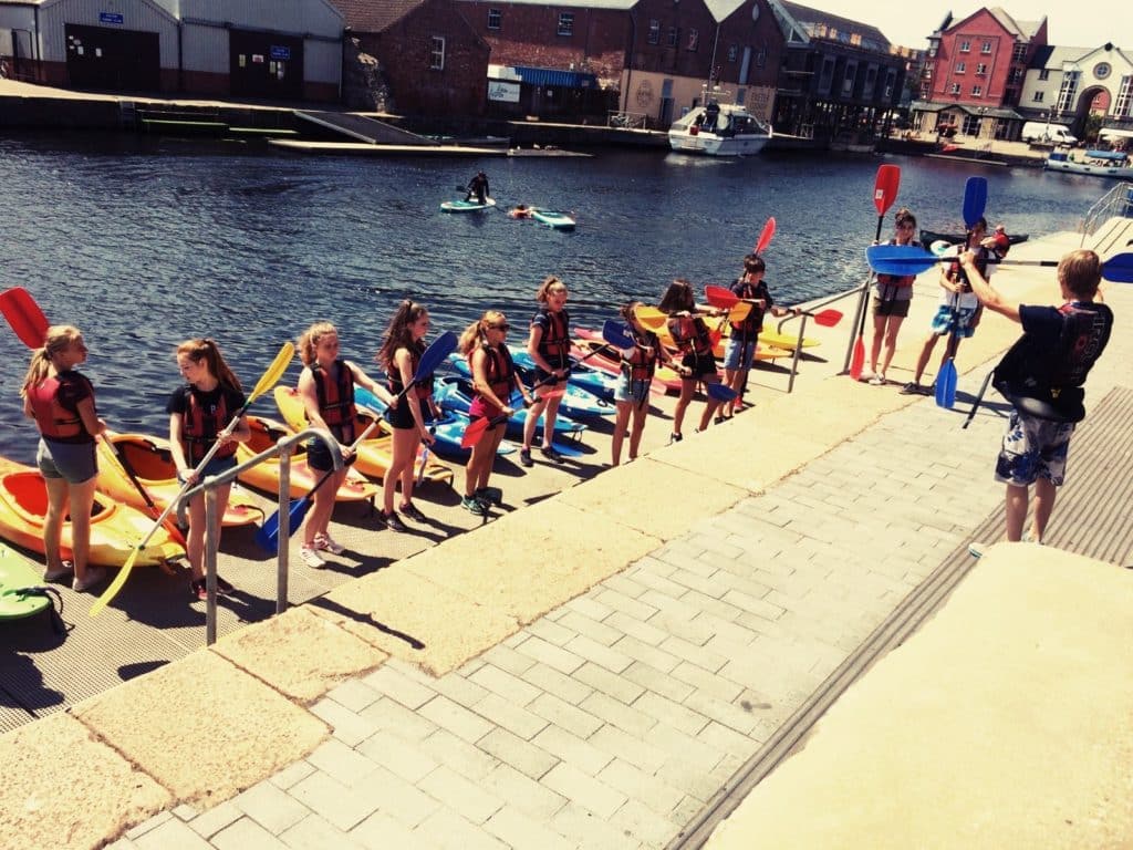 English students learning how to canoe. Students are standing holding their oars on the platform looking at the instructor. The canal is behind them